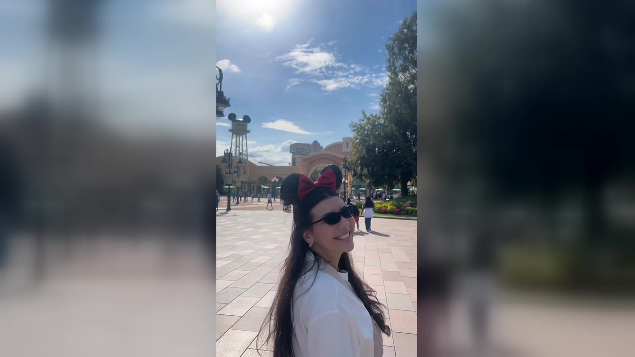 Karol Barbosa de orelhinha da Minnie sorrindo na praça de entrada da Disneyland Paris, com a caixa-d’água do Mickey ao fundo